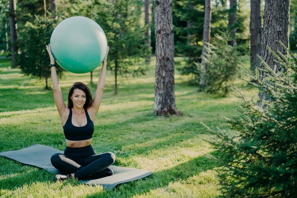 Brunette slim woman lifts fitness ball over head, sits in lotus pose on karemat, dressed in active wear, practices yoga outside, breathes fresh air in forest. Fit lady exercises with gymnastic ball