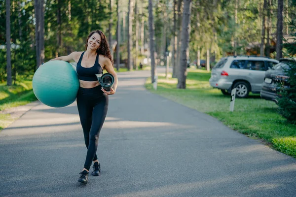 Full length shot of active slim woman carries fitness ball and rolled up karemat, dressed in leggings, top and sneakers, walks on path near park or forest, looks gladfully, has athletic body