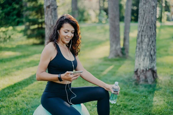 Pleased relaxed woman has rest after yoga exercises sits on fitness ball, listens music via modern mobile phone and earphones, holds bottle of fresh water, dressed in active wear, poses at park