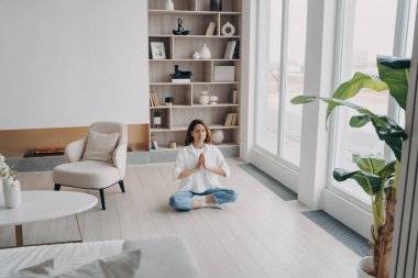 Huzurlu kadın yoga yapıyor modern oturma odasında yerde oturuyor namaste el hareketi yapıyor. Sakin bir kadın içeride temiz hava soluyor, meditasyon yapıyor. Farkındalık, stres atma kavramı.