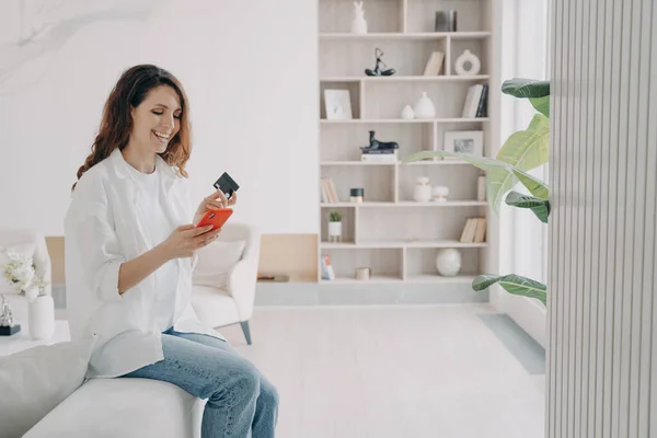 Smiling hispanic female holding bank credit card, smartphone, making successful payment, shopping in online store, sitting in living room at home. E commerce, e-bank apps, banking services advertising