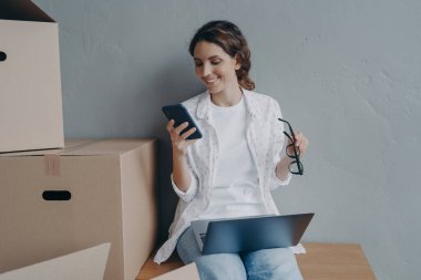 Mutlu iş kadını cep telefonunda uygulama kullanıyor, paket siparişi veriyor. Bayan ofis müdürü, çevrimiçi sipariş kutularını göndermek için hazırlıyor. E-ticaret, küçük işletme konsepti.