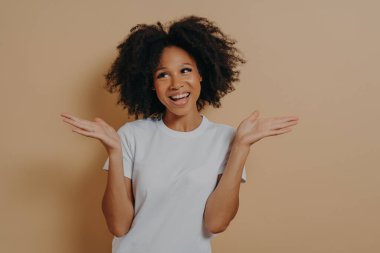 Genç, gülümseyen kıvırcık ırklı kadın iki elini de tereddütle kaldırıyor ve yan bakıyor, karar veriyor ya da beyaz tişörtlü, bej arka planda izole edilmiş bir şey seçiyor.