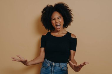 Siyah tişörtlü Afrikalı kadın çığlık atıyor, avuçlarını açıyor, kızgın hissediyor, ağzı ardına kadar açık kameraya bakarak bağırıyor, bej renkli stüdyo arka planında izole edilmiş. Olumsuz duygular konsepti