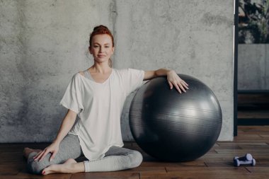 Güzel, sportif, kızıl saçlı bir kadının portresi ahşap zeminde spor salonunda ya da pilates stüdyosunda oturur ve kameraya mutlu bir şekilde gülümserken bir eliyle halterin yanına uzanır.