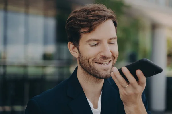 Portrait of handsome smiling bearded office worker talking on speakerphone, recording audio message, using virtual digital voice assistant, standing outside of mirrored business center building