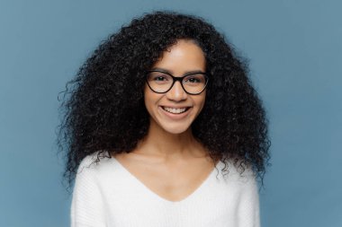 Hoş bir gülümseme ile güzel kıvırcık kadın Kapalı çekim, gözlük ve beyaz rahat jumper giyer, mavi arka plan üzerinde izole, iyi duygular ifade eder. Kadınsı kız arkadaşları ile güzel bir gün sahiptir