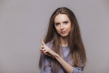 Boş kopya alanı gri duvar, noktalarda esmer uzun saçlı sevimli genç kadın modeli ciddi ifade ile bir şey advertizes, dikkatinizi çekiyor. Reklam ve insanlar kavramı
