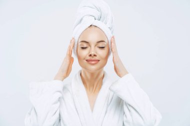 Güzel spa kadını kapalı gözlerle ayakta duruyor, ellerini kafaya sarıp beyaz banyo bornozu giyiyor, sağlıklı cilde, doğal makyaja sahip, bakımlı tene sahip, içeride poz veriyor. Güzellik tedavisi