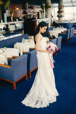 Çekici ince genç esmer gelin kapalı atış harika beyaz gelinlik giyer, Festival salonunda Konuklar için bekler, ifade korkar mı, dikkatle buket görünüyor, geri duruyor