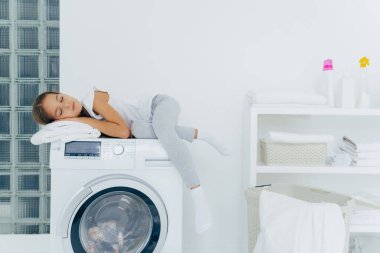Horizontal shot of little girl has rest on washing machine, feels tired after helping mum about house, sleeps on washer poses in laundry room with console, detergents, linen around. Household routine
