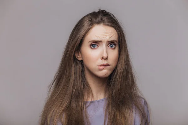 Offended stressful female bites lower lip with dissatisfaction, has sorrorful expression, expresses negative emotion, being in low spirit after quarrel with somebody, isolated over grey background