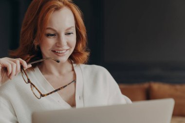 Güzel kızıl saçlı kadın dizüstü bilgisayar ekranına odaklanmış, olumlu gülümsüyor, optik gözlük tutuyor, içeride poz veriyor, modern teknolojileri online iletişim için kullanıyor, Webinar izliyor, haber okuyor.