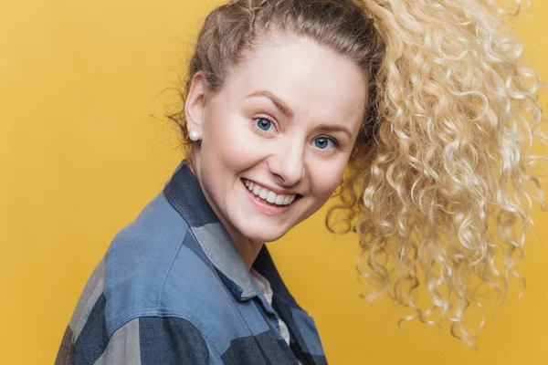Sideways shot of pleased adorable young female model with curly hair, broad shining smile, enjoys spare time with close people, happy to have day off, isolated over yellow background. Positiveness