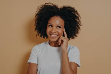 Aklınızda bulunsun. Dalgın, genç bir Afrikalı kadının portresi kıvırcık saçlı, işaret parmağıyla şakağa dokunan, neşeli gülümseyen, iyi bir fikri olan, bej arka planda izole edilmiş poz veren.