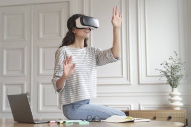 Sanal gerçeklikte bir şeye dokunmak isterken evde uzaktan çalışırken bilgisayarla serbest çalışma masasında oturan genç, şaşkın kadın çalışan VR kulaklık takıyor.