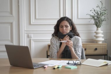 Grip olmuş. Hasta genç İspanyol kadın ofis çalışanı iş yerinde termometre tutuyor ve vücut ısısını ölçüyor. Hasta kadın çalışanın kovid 19 koronavirüs belirtileri var.