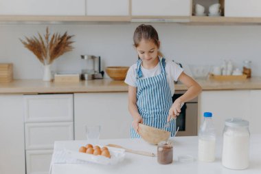 Şirin küçük kız çizgili önlük giyiyor, malzemeleri kaseye çırpıyor, hamur hazırlıyor, yemek yapmayı öğretiyor, yumurta, süt ve un ile mutfak arka planında beyaz masada duruyor. Childern ve aşçılık konsepti