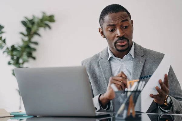 Puzzled frowning african american businessman discussing working project online by video call at laptop. Pensive dissatisfied black male manager solving work problem, holding paper report.