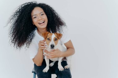 Köpeğini okşayan neşeli kıvırcık kızın portresi, Jack Russell terrier almaktan mutlu, geniş bir şekilde gülümseyen, hayvanlarla oynayan, günlük kıyafetler giyen, beyaz arka planda izole edilmiş, iyi bir günün tadını çıkaran