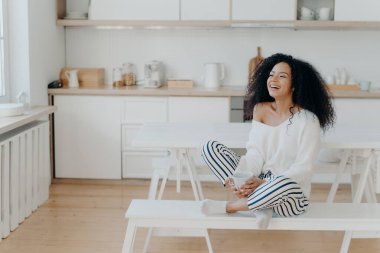 Aşırı neşeli, koyu tenli bir kadının hoş bir şekilde kahkaha attığı, kahve içtiği, mutfaktan dışarı bakıp şık kıyafetler giydiği yatay bir sahne. Sıcak bir içecekle gülümseyen bayan, evde dinleniyor.