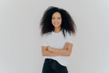 Afro saç stili, kollarını kavuşturmuş, neşeli bir şekilde gülümseyen, beyaz arka planda izole edilmiş günlük tişört ve kot pantolon giyen kaygısız genç bir kadının stüdyo fotoğrafı. İnsanlar, etnik köken, yüz ifadeleri