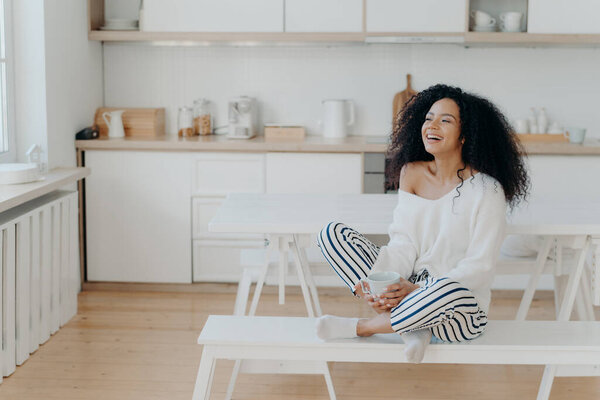Горизонтальный кадр радостной темнокожей женщины приятно смеется, пьет кофе, смотрит в окно на кухне, одевается в модную одежду. Улыбающаяся леди с горячим вкусным напитком, расслабляется дома
