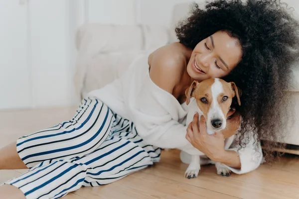 愛情の若い女性は愛とケアで犬を抱擁し 目を喜びから閉じ 優しく笑顔を維持し 床の上に健康な暗い肌 ポーズ ペット動物があります 人と友情とペットの概念 — ストック写真