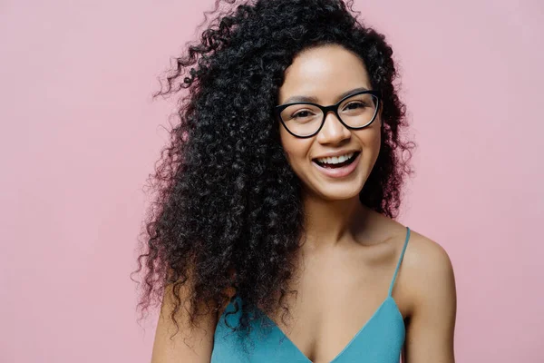 Close up shot of happy carefree African American woman laughs from joy, has healthy dark skin, curly hair, isolated over pastel rosy background, hears compliment. People, emotions, happiness concept