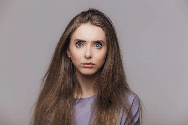 Studio shot of pleasant looking blue eyed female model with dark long hair, has puzzled expression as hears shocking news from interlocutor, isolated over grey background. Facial expressions concept