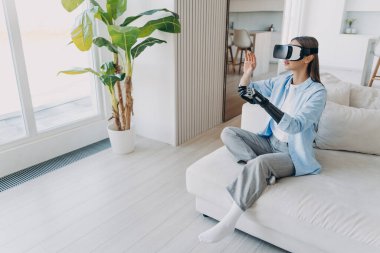 Özürlü, VR gözlüklü, yapay eli hareket ettiren, kanepede oturan modern bir kız. Engelli genç kadın duyusal biyonik protez kol kullanarak sanal gerçeklikle etkileşime geçerek rehabilitasyon görüyor..