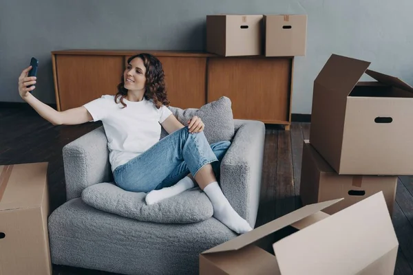 Smiling girl holding smartphone, chatting by video call in new apartment during relocation. Female communicates with friends or relatives, showing her house with cardboard boxes on moving day.