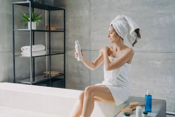 Young woman applies cream to her shoulder. Body lotion applying. Attractive caucasian girl wrapped in towel after bathing. Cosmetic products for smooth silky skin. Spa procedure at home.