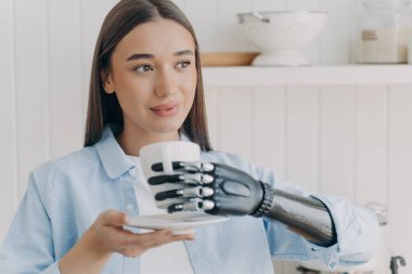 Özürlü kızın elinde siber ellerle bir fincan çay tutuyor. Mutfaktaki mutlu engelli kız. Makyajlı güzel kadın gülümsüyor. Modern elektronik protezdeki algılayıcı kavramı.