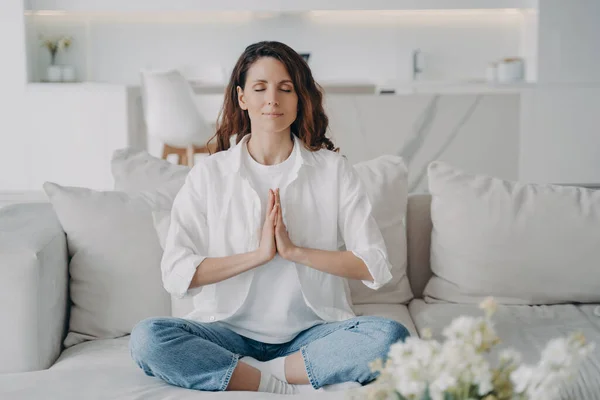 Peaceful spanish woman relaxing in lotus pose on comfortable couch at home. Calm female enjoying practicing yoga breathing exercises, meditation, sitting on sofa. Reducing stress, wellness concept.