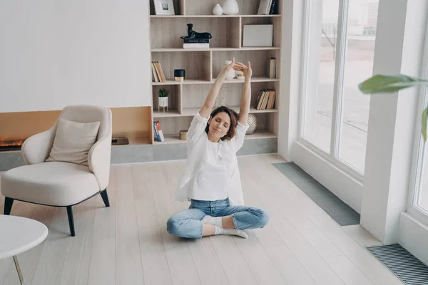 Calm female sitting in lotos position on floor in living room, stretching arms up. Woman practicing yoga, taking a break, doing antistress exercises at home. Healthy lifestyle, wellness concept.