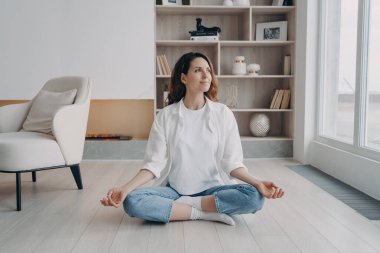 Barışçıl İspanyol kadın Padmasana 'da yoga yapıyor, lotus poz veriyor oturma odasında. Sakin bir kadın meditasyon yapıyor, evde nefes egzersizleri yapıyor. Stres atma, sağlıklı yaşam tarzı konsepti.