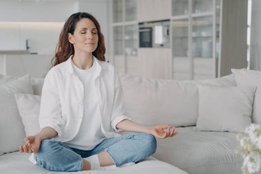 Sakin bir kadın meditasyondan zevk alıyor, kanepede nilüfer pozisyonunda yoga yapıyor. İspanyol kadın dinleniyor, evdeki kanepede nefes egzersizleri yapıyor. Sağlıklı alışkanlıklar, düşünceli yaşam tarzı, endişe giderme.