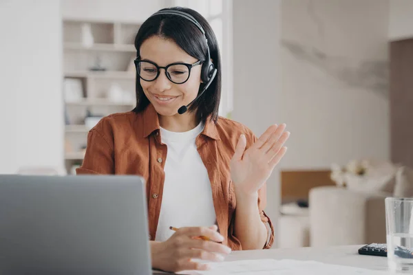 Smiling female business coach in glasses, headset, conducts online consultation by video call, greeting client. Businesswoman waving hand hello, talking with coworkers on distance corporate meeting.