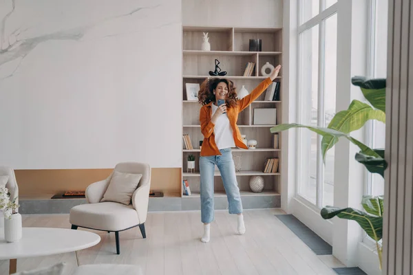 Happy dancing young girl holding smartphone, listening to music by mobile apps, moving to song in modern living room. Energetic female dances to musical playlist, rejoicing with good news at home.