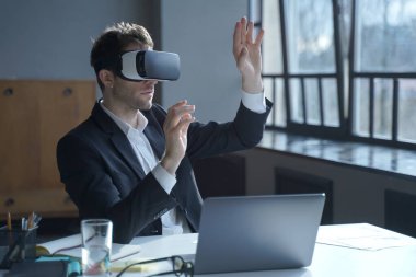 VR kulaklıklı bir erkek ofis çalışanı masada otururken dijital arayüzle etkileşime giriyor, havada üç boyutlu nesnelere dokunuyor, takım elbiseli bir işadamı sanal gerçeklik gözlüklerini iş dünyası için test ediyor