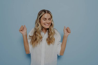 Heyecanlı, sevinçle dolu, iki eli havada tezahürat yapan genç sarışın kız mavi duvarın üzerinde soyutlanmış bir şekilde poz verirken duygularını jestlerle ifade ederek mutluluğunu kontrol edemiyor. Heyecan konsepti