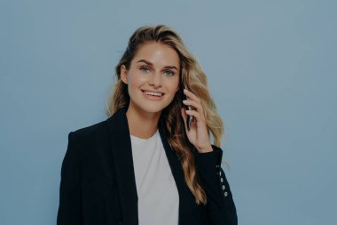 Kendine güvenen genç ofis çalışanı cep telefonuyla konuşuyor, kameraya bakıyor, mavi arka planda tek başına duruyor, beyaz gömleğin üzerine siyah ceket giyiyor.