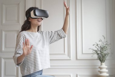E-Ticaret 'te VR. Sanal gerçeklik kulaklıklarıyla evde dikilen ve gelişmiş dünyada eğlenceli alışveriş deneyimlerinden zevk alan, ellerle nesnelere dokunan, gündelik kıyafetlerle mutlu genç bir kadın.