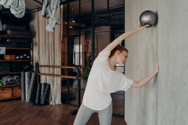 Genç, beyaz tenli, spor salonundaki spor salonunda küçük topa uygun spor yapan, etkin tişört ve tozluk giyen kızıl saçlı kadın. Spor ve pilates konsepti