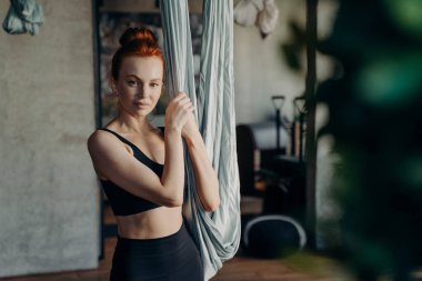 Sağlıklı, kızıl saçlı genç bir kadın spor salonunda duruyor ve gri hamağı tutuyor. Sinek fitness antrenmanı sırasında kameraya bakıyor. Yerçekimi yogasına hazırlanıyor.