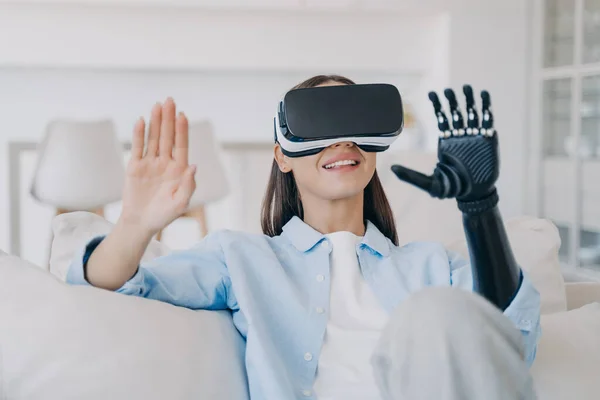 Smiling young girl wearing virtual reality glasses learning to use bionic prosthetic arm after limb loss. Disabled woman in vr goggles testing artificial hand, myoelectric prosthesis.