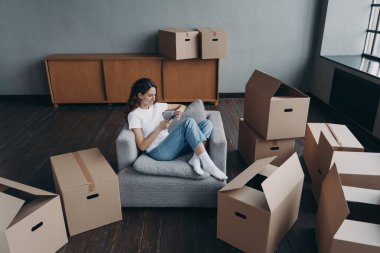 Genç bir kadın online cep telefonu uygulaması kullanıyor, mobilya alıyor taşınma gününde karton kutuların arasında oturuyor. Paketlenmiş şeyleri yeni eve taşımak için taşınma servisini seçen kadın.