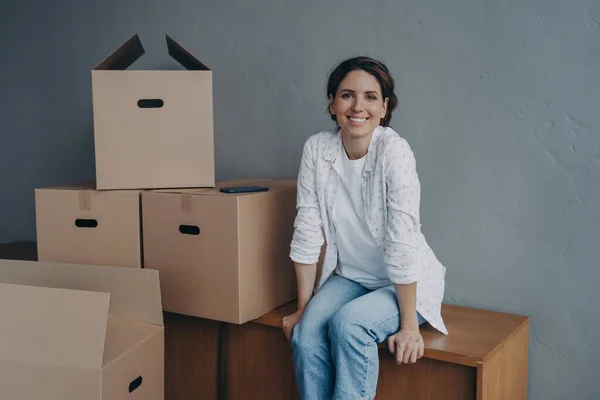 Smiling hispanic woman sitting with cardboard boxes in new rent house or apartment. Mid adult female satisfied with preparation of carton parcels on moving day at home. Real estate, relocation concept