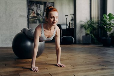 Genç, gülümseyen, kızıl saçlı bir kadın spor salonunda antrenman yapıyor, spor topunun üzerine uzanıyor ve kollarını yere dayayıp şınav çekiyor sportif güdümlü bir kadın Fitball 'la çalışıyor.